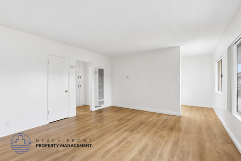 A spacious room with wooden flooring and white walls, with a door on the left and a window on the right.