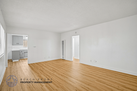 A spacious room with wooden flooring and white walls, with a logo for Beach Front Property Management in the bottom left corner.