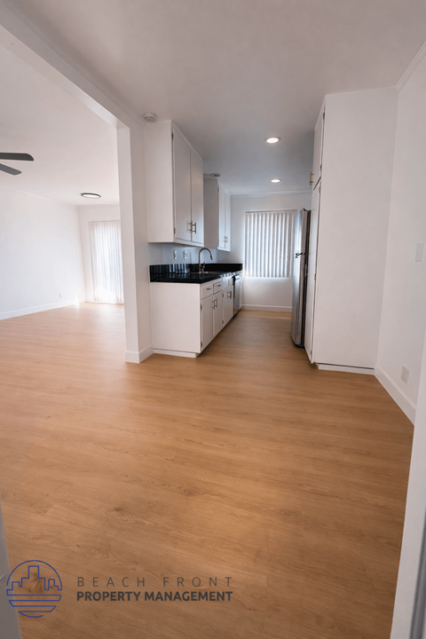 A spacious, empty room with wooden floors and white walls, with a kitchen area visible in the background.