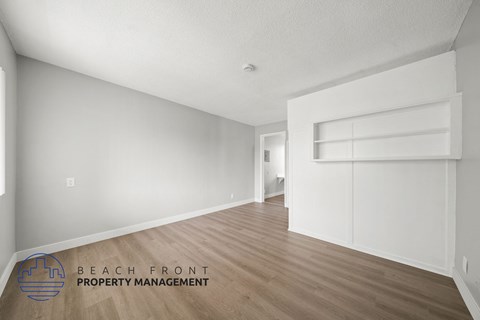 A spacious room with wooden flooring and white walls, with a logo for Beach Front Property Management in the bottom left corner.