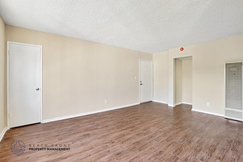 A spacious room with wooden flooring and white walls, with a door on the left and a window with a screen on the right.