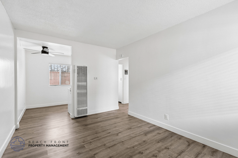 A spacious room with wooden floors and white walls, featuring a ceiling fan and a window with blinds.