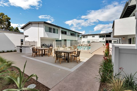 A patio with a table and chairs is surrounded by plants and a pool is in the background.