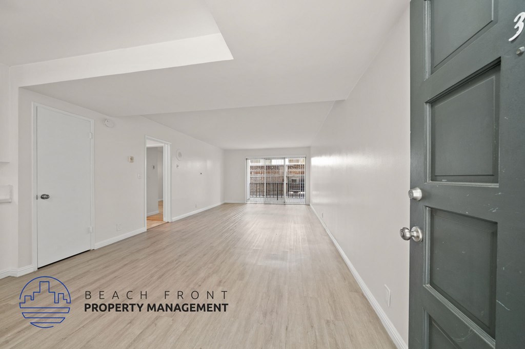 A spacious hallway with wooden floors and white walls, leading to a kitchen area with a sink and stove.