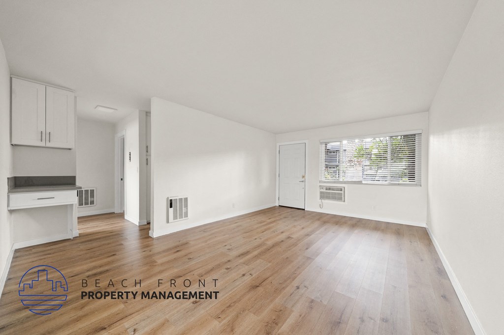 A spacious room with wooden flooring and white walls, with a logo for Beach Front Property Management in the bottom left corner.