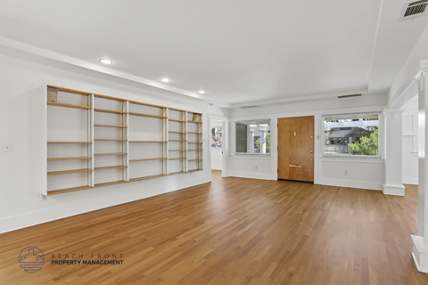 A spacious room with wooden floors and white walls, featuring a large window with blinds and a wooden door.