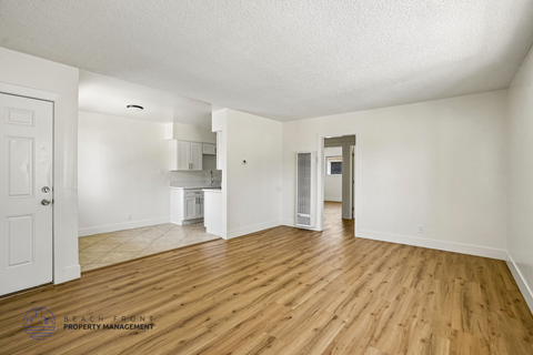 A spacious room with wooden floors and white walls, with a door on the left and a kitchen area visible in the distance.