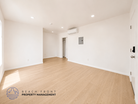 A spacious room with wooden flooring and white walls, with a logo for Beach Front Property Management in the bottom left corner.