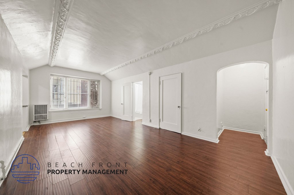 A spacious room with wooden floors and white walls, with a window and a door visible.