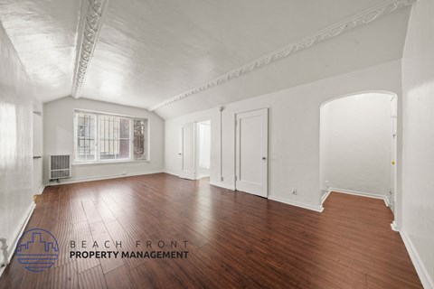 A spacious room with wooden floors and white walls, with a window and a door visible.