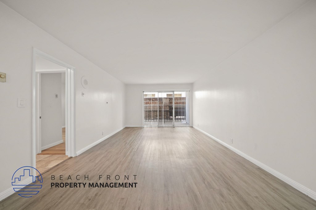 A spacious room with wooden flooring and white walls, with a balcony visible through the window.