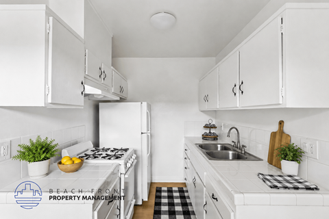 A kitchen with white cabinets and appliances, a black and white checkered mat on the floor, and a logo for Beach Front Property Management in the corner.