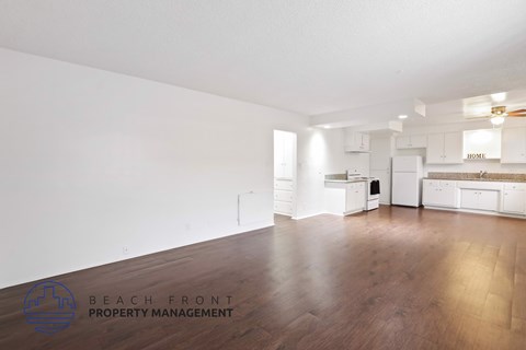A spacious, empty room with wooden floors and white walls, with a logo for Beach Front Property Management in the bottom left corner.
