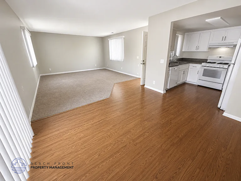 A spacious living room with wooden floors and a kitchen area in the background.