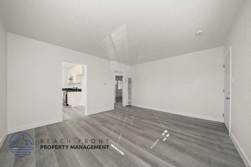 A spacious room with wooden flooring and white walls, with a logo for Beach Front Property Management in the bottom left corner.