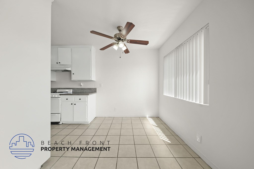 A room with a ceiling fan and a kitchen area in the background.