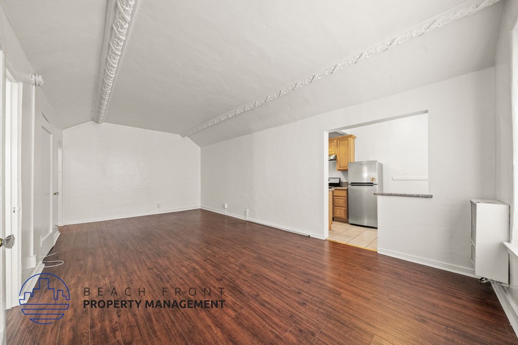 A spacious room with wooden floors and white walls, with a kitchenette visible in the background.
