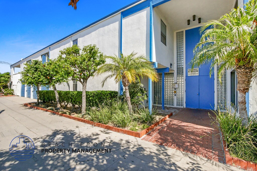 A blue building with a white door and windows, surrounded by greenery and a red brick pathway.