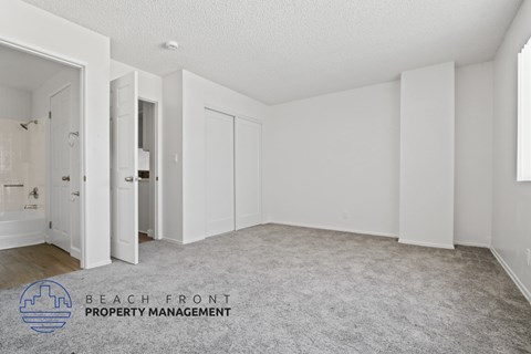 A spacious, empty room with a carpeted floor and white walls, with a logo for Beach Front Property Management in the bottom left corner.