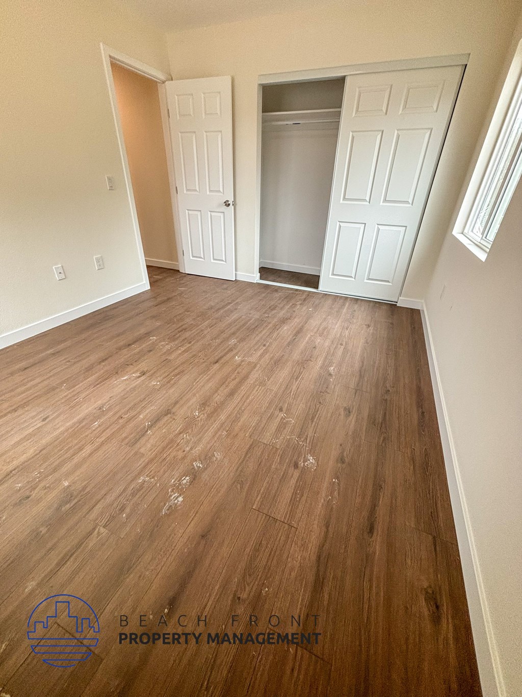 A room with wooden floors and white walls, with a logo for Beachfront Property Management in the bottom left corner.
