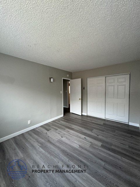 A room with a wooden floor and white walls, with a logo for Beach Front Property Management in the bottom left corner.