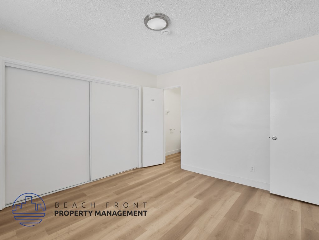 A room with white walls and wooden flooring, with a logo for Beach Front Property Management in the bottom left corner.