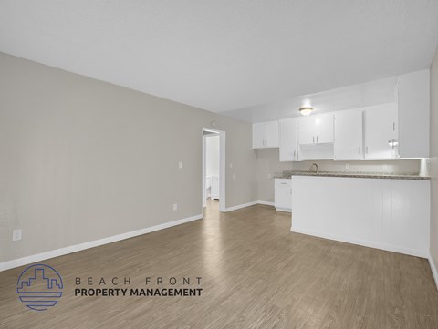 A spacious room with wooden flooring and white walls, featuring a kitchen area with white cabinets and a countertop.