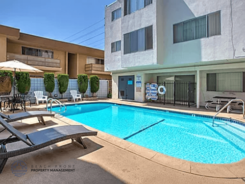 A pool surrounded by a patio with chairs and a building in the background.