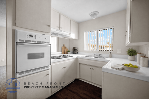 A kitchen with white cabinets and appliances, a window with blinds, and a bowl of fruit on the counter.