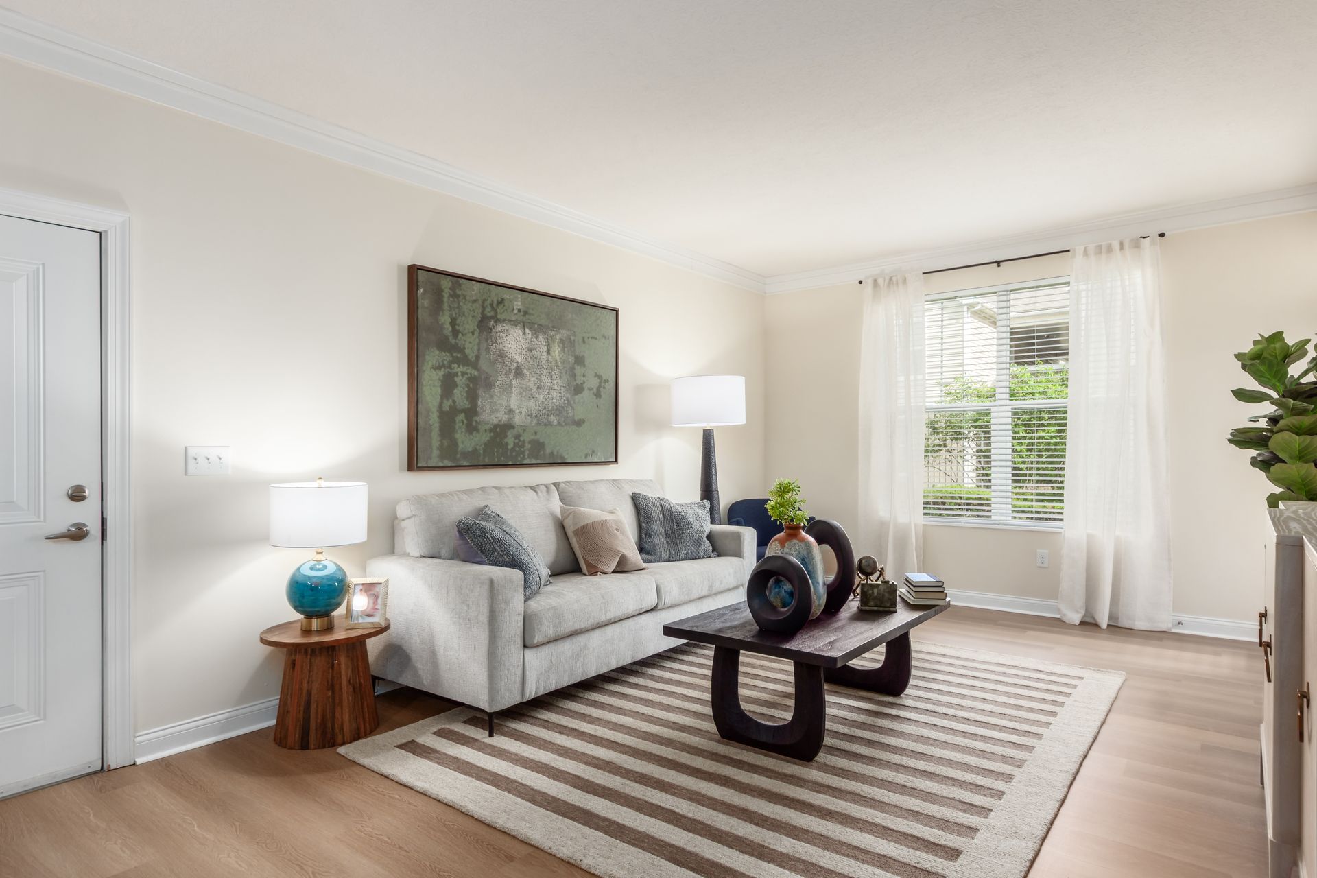 A living room with a grey couch, a wooden side table with a blue vase, a coffee table, and a painting on the wall.