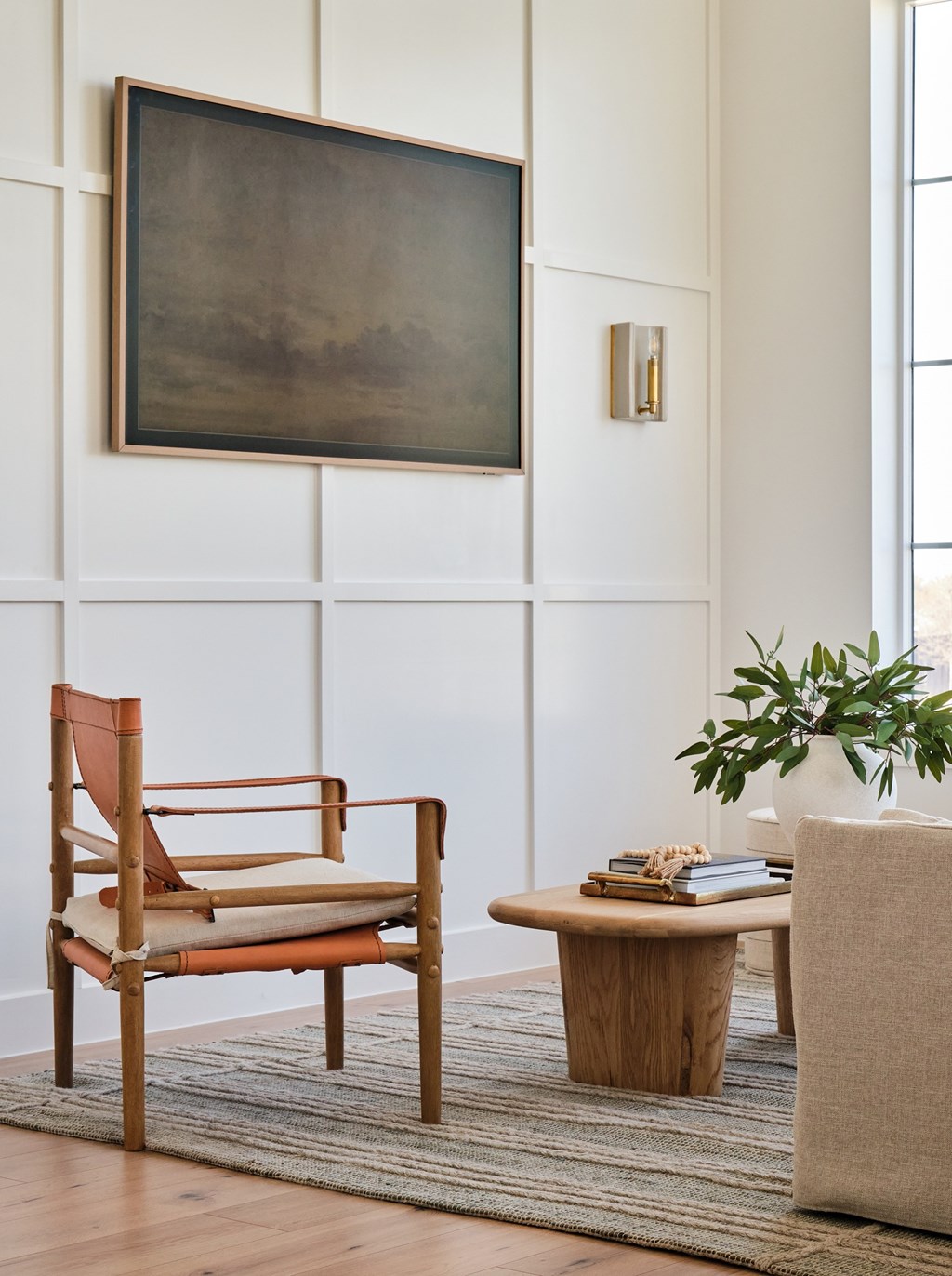 A chair with a cushion sits in front of a wall with a painting and a small table with a plant on it.