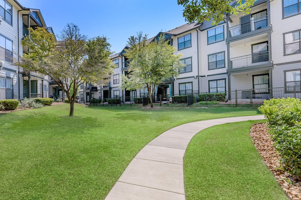 a sidewalk leading to an apartment building with green grass and trees