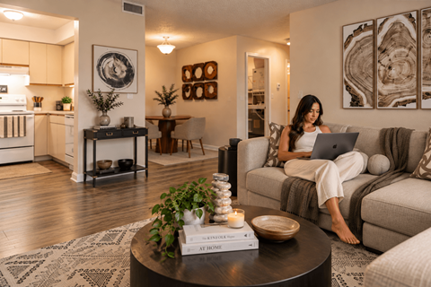 A woman is sitting on a couch with a laptop in a well-lit living room.