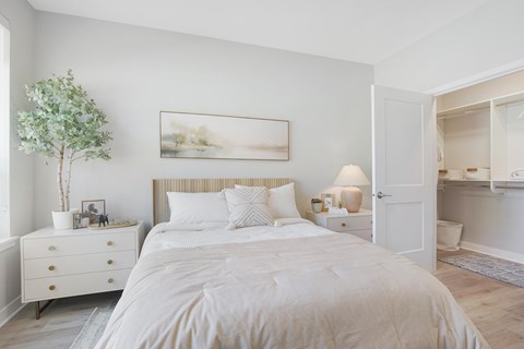 A white bedroom with a large bed, a nightstand with a lamp, and a painting on the wall.