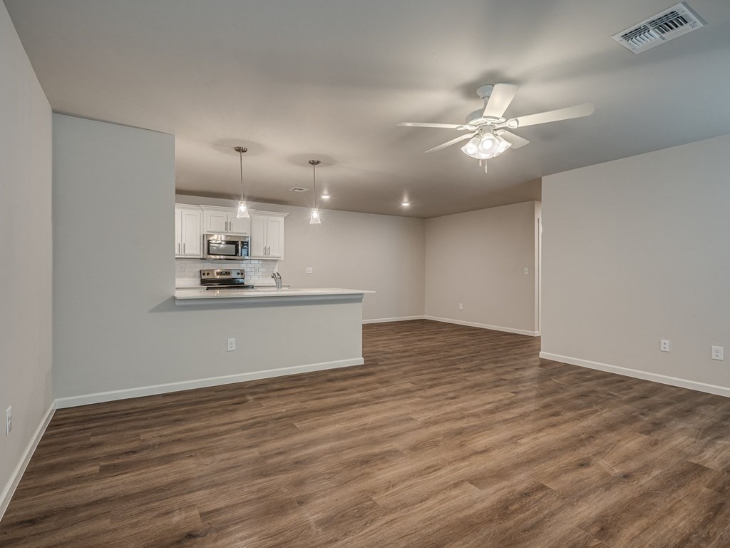 an empty living room with a ceiling fan and a kitchen in the background