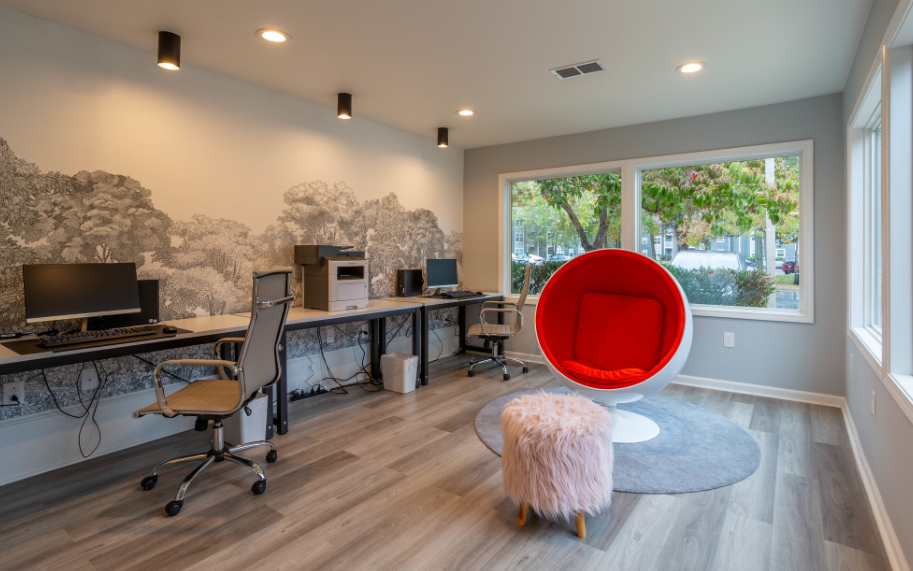 a bright red egg chair sits in front of two computers in an office with a large window