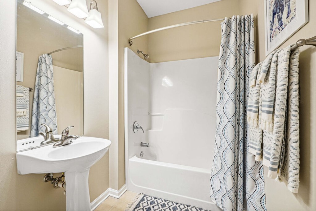 A white sink is on the left of a bathroom with a white tub and a white towel hanging on the towel rack.