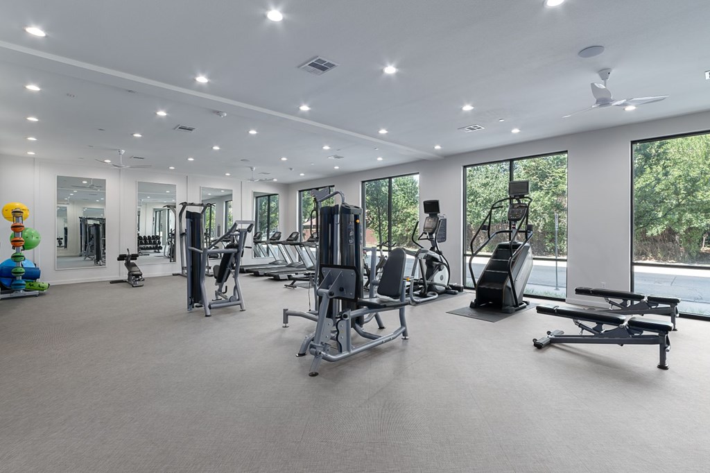 A spacious gym with a variety of equipment and a view of trees outside the windows.
