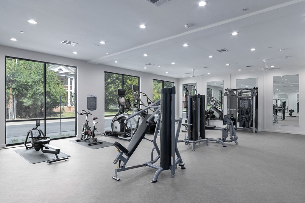 A spacious gym with a variety of equipment and a view of trees outside the windows.