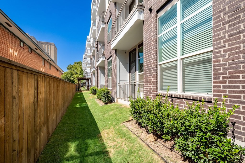 A long, narrow, green lawn separates a brick building from a wooden fence.