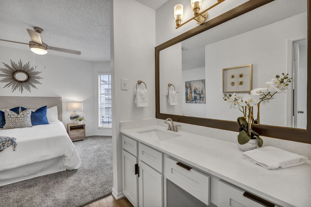A well-lit, white bathroom with a large mirror and a bed in the background.