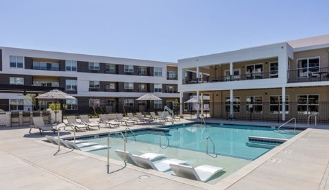 A large swimming pool surrounded by lounge chairs in front of apartment buildings.