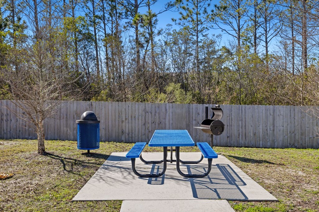 A blue picnic table is in the middle of a concrete slab with a blue trash can and a tree in the background.