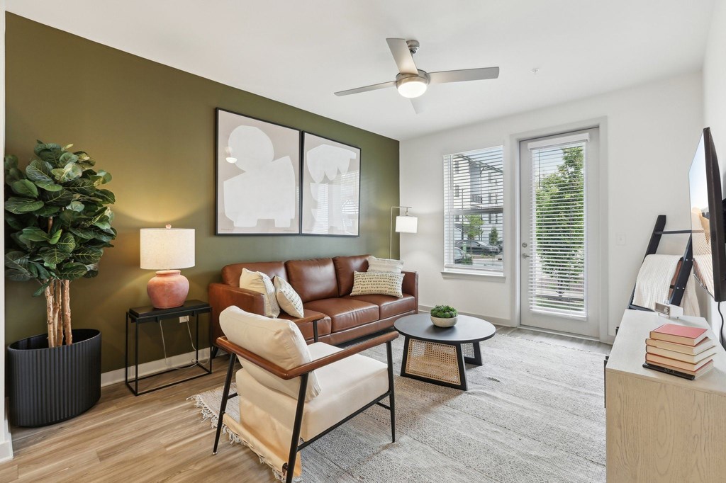 A living room with a brown leather couch, a white chair, a small table, a potted plant, and a ceiling fan.