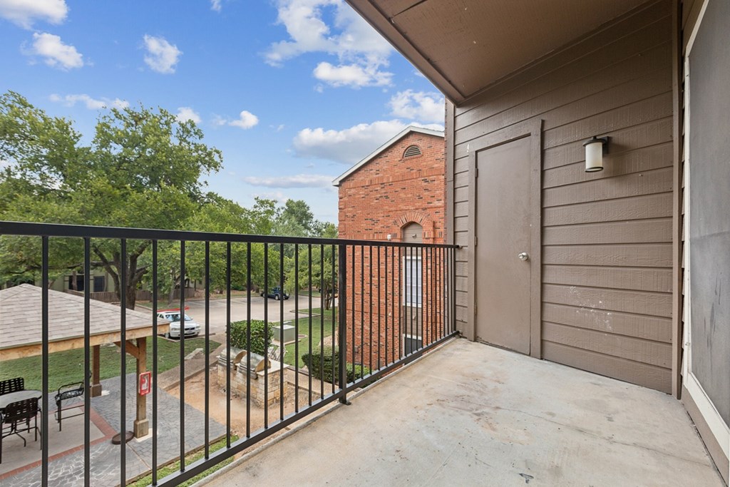 A balcony with a black railing overlooks a courtyard with a brick building in the background.