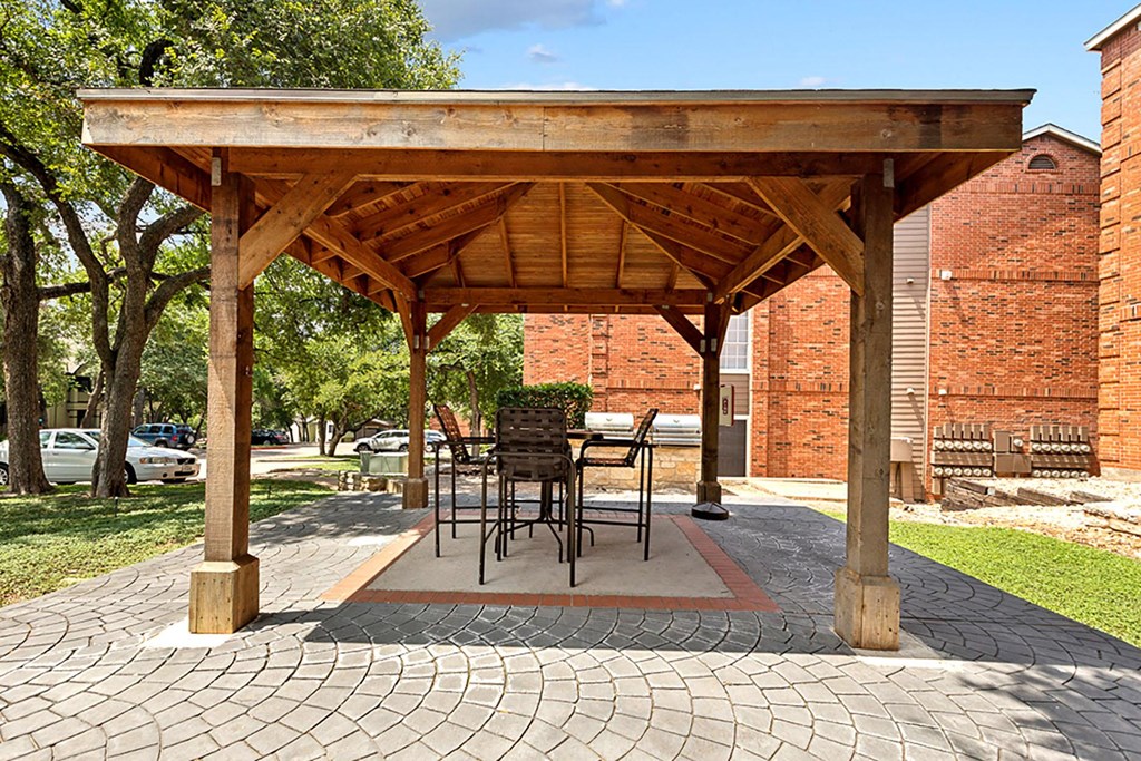 A wooden pavilion with a table and chairs is surrounded by brick buildings and trees.