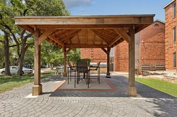 A wooden pavilion with a table and chairs is surrounded by brick buildings.