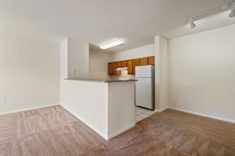an empty living room with a kitchen with a refrigerator