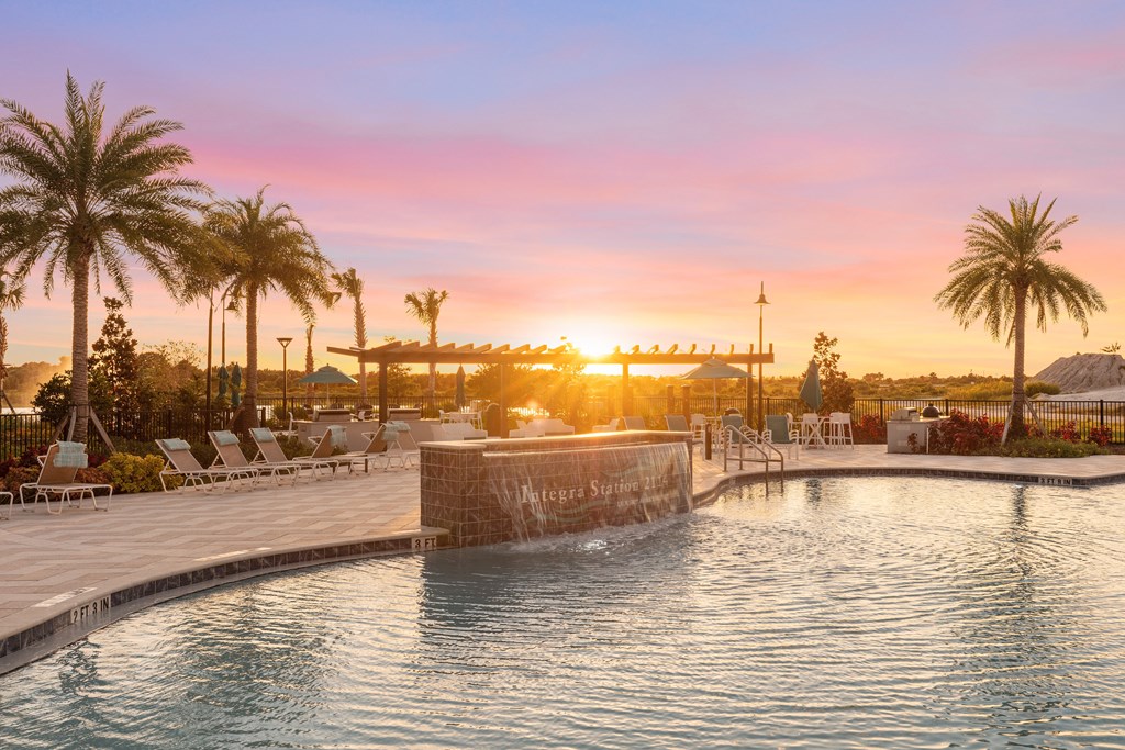 A sunset view of a pool with a waterfall and palm trees.