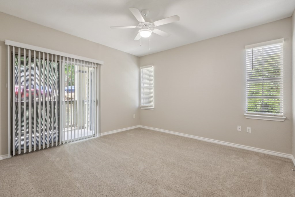 A room with a fan, carpeted floor, and a window with blinds.